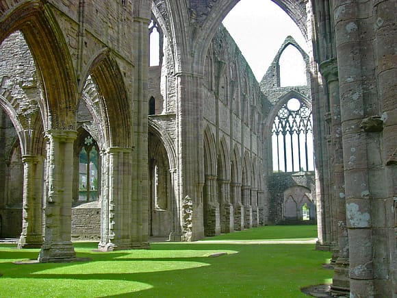 Photograph of the nave and stonework of Tintern Abbey, now in ruins, but with tall walls still standing with arched side chapels and spaces. The ground is a beautiful flat carpet of green grass.