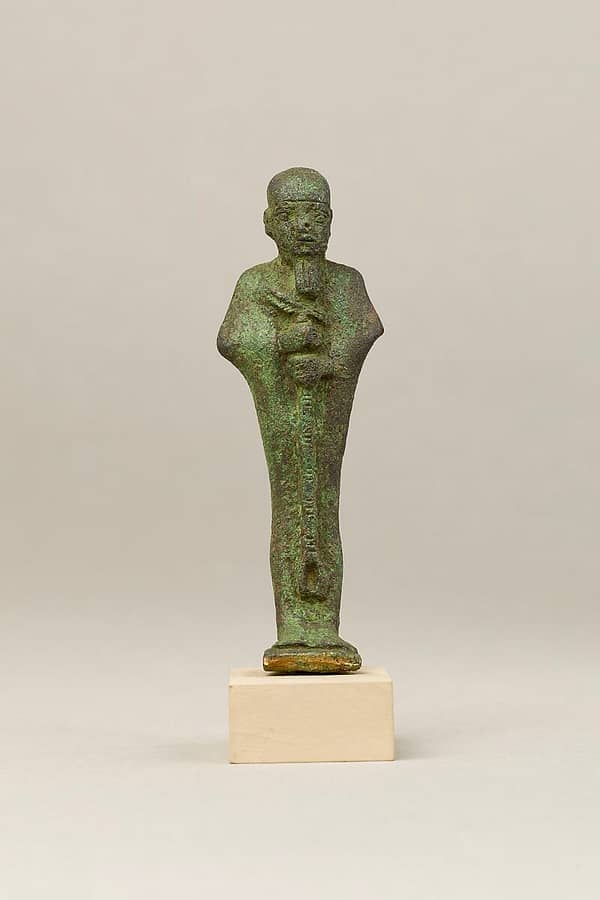 A metal statue of the god Ptah, with somewhat unbalanced facial features and unevenly carved eyes, wearing the tight-fitting cap, straight beard, and carrying a scepter.